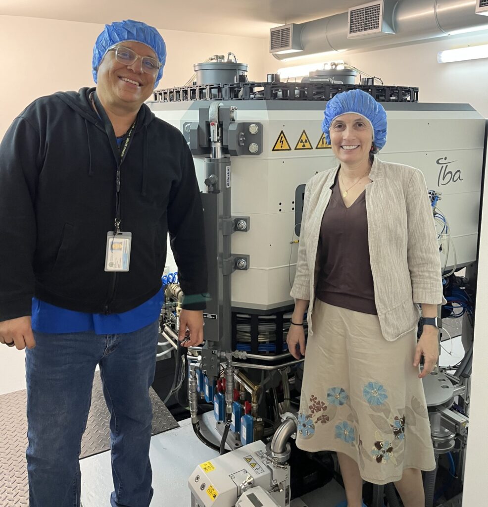 Two people smile in front of large machine with hairnets on.